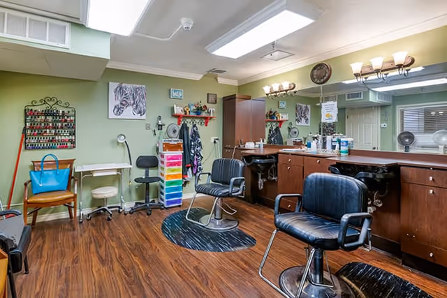 Interior view of a salon area in a senior living facility with two black salon chairs, a wooden floor, a large mirror with lights above it, a colorful storage drawer unit, nail polish racks on the wall, and various salon tools and decorations.