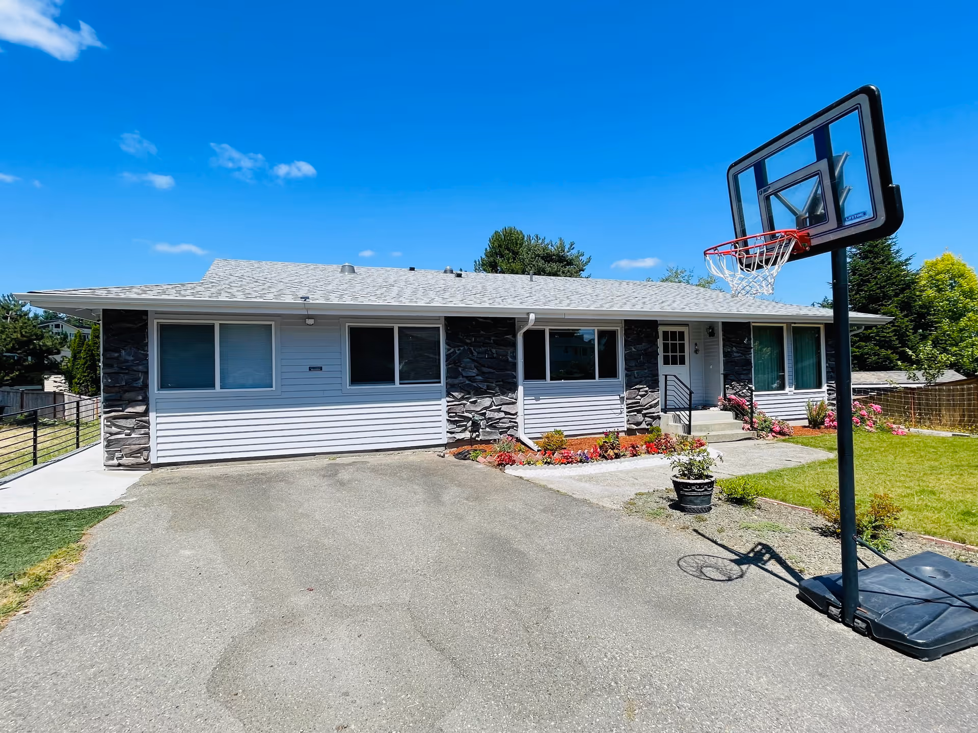 Single-story residential building with white siding and stone accents, a small garden with flowers near the entrance, a paved driveway, and a portable basketball hoop on the right side under a clear blue sky.