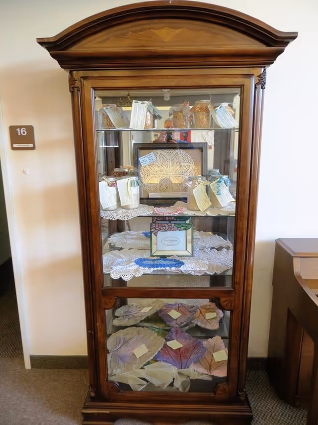 A wooden display cabinet with glass doors containing various handcrafted items including jars with tags, a framed lace piece, and colorful leaf-shaped crafts. The cabinet is placed against a wall next to a door labeled with the number 16.
