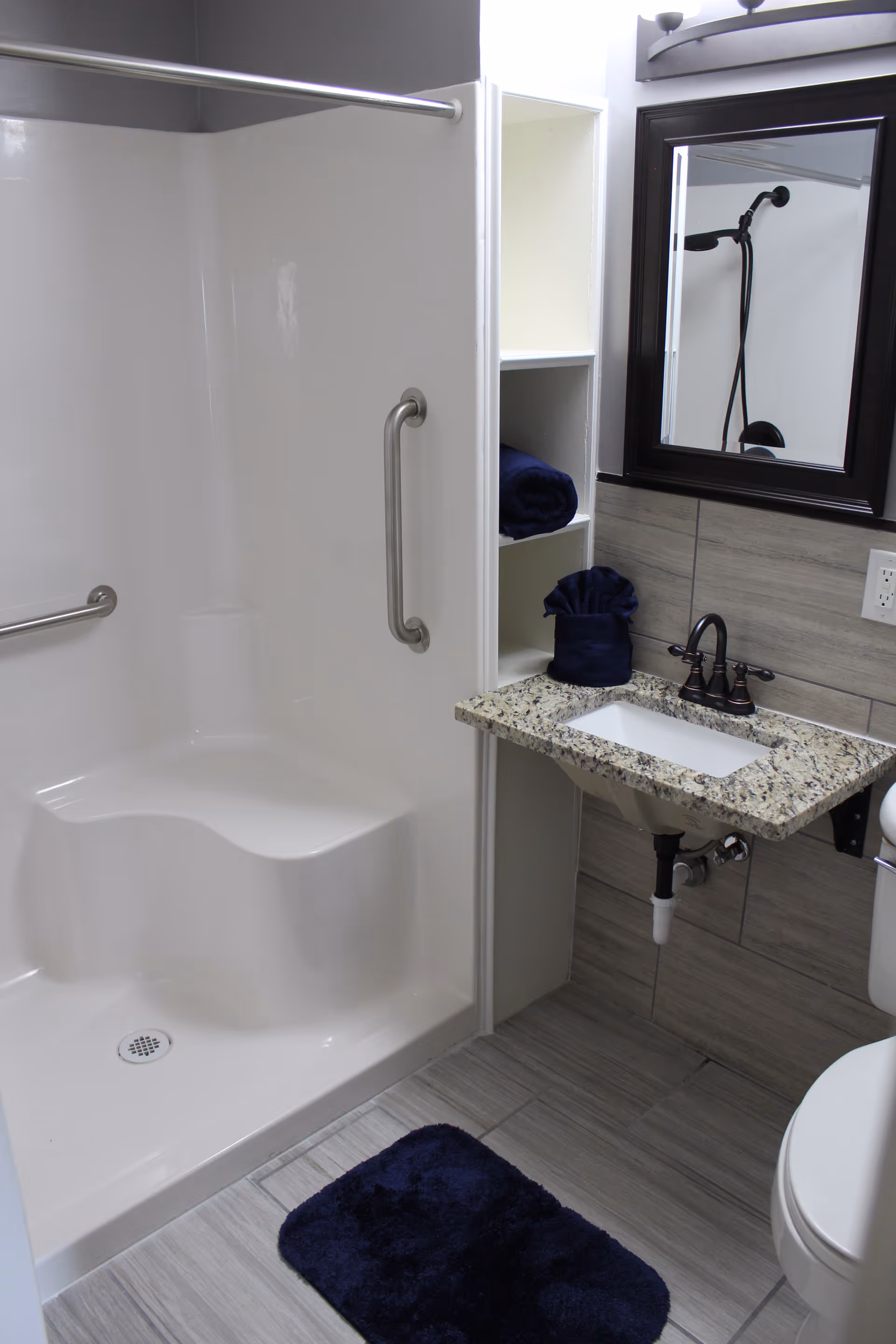 A clean and modern bathroom featuring a white walk-in shower with built-in seat and grab bars, a granite countertop sink with dark faucet, a mirror above the sink, open shelving with rolled dark blue towels, a dark blue bath mat on the tiled floor, and a partial view of a white toilet.