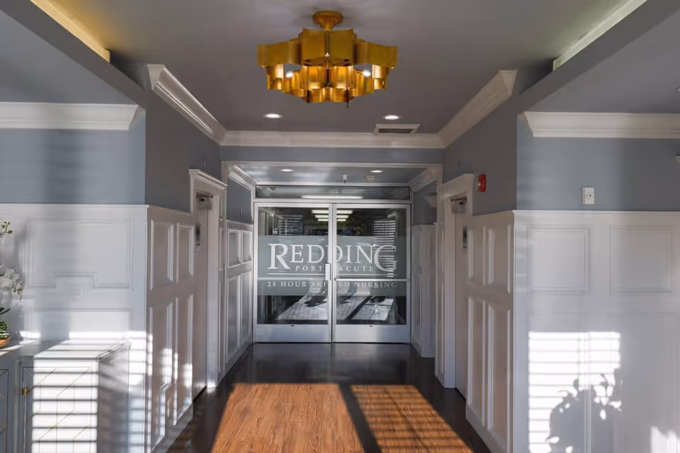 Interior hallway with wood flooring and white wainscoting leading to glass entrance doors bearing the text 'Redding Post Acute'.