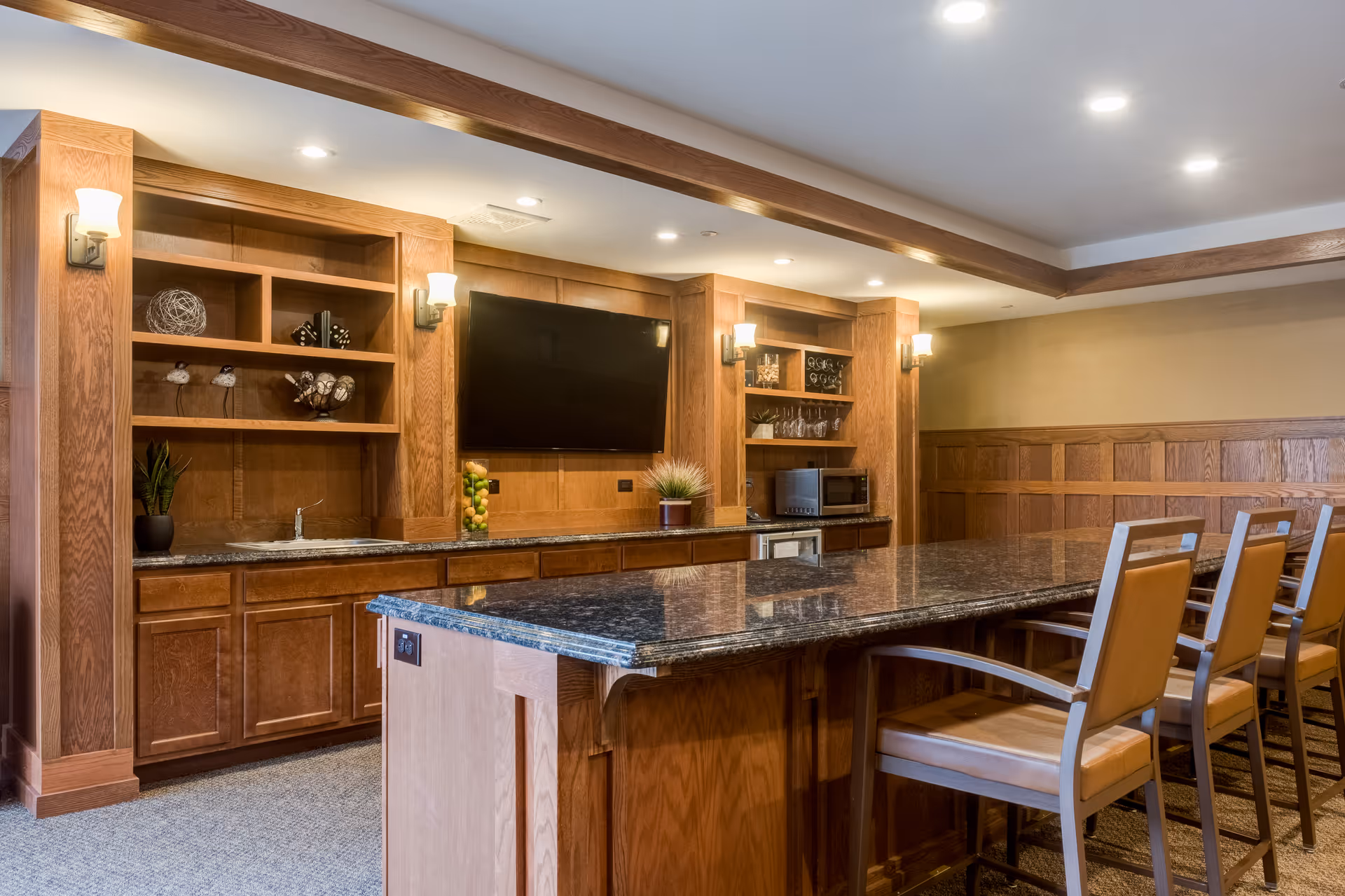 A spacious interior room with wooden cabinetry and paneling, featuring a large granite countertop island with four high-backed chairs. The back wall has built-in shelves with decorative items, a mounted flat-screen TV, a small sink, and a microwave. The room is well-lit with recessed ceiling lights and wall sconces.