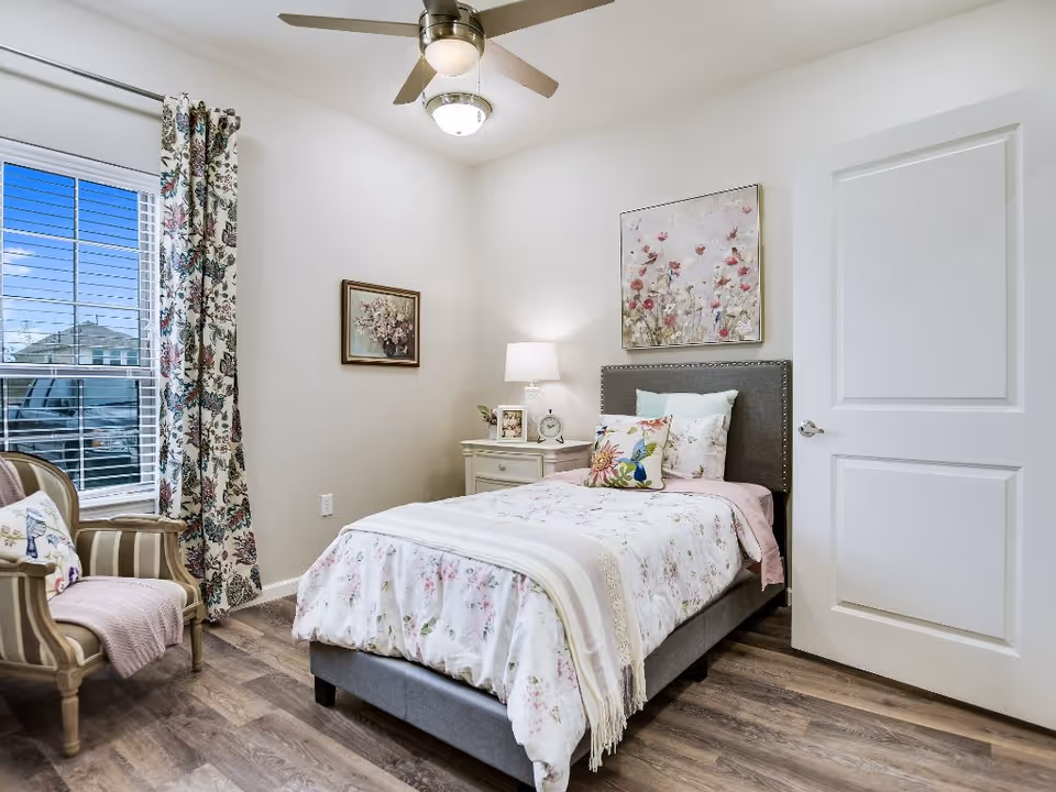 Bright, neatly furnished bedroom featuring a single bed with floral bedding, a nightstand and lamp, an armchair by a window, and wall art.