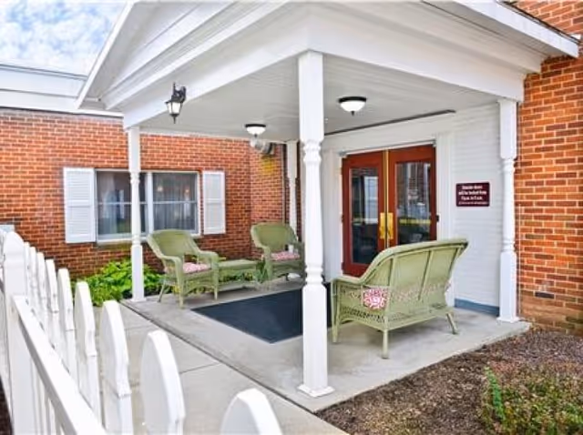 Covered outdoor seating area with green wicker chairs and a loveseat with cushions, located at the entrance of a brick building with white trim and double red doors.