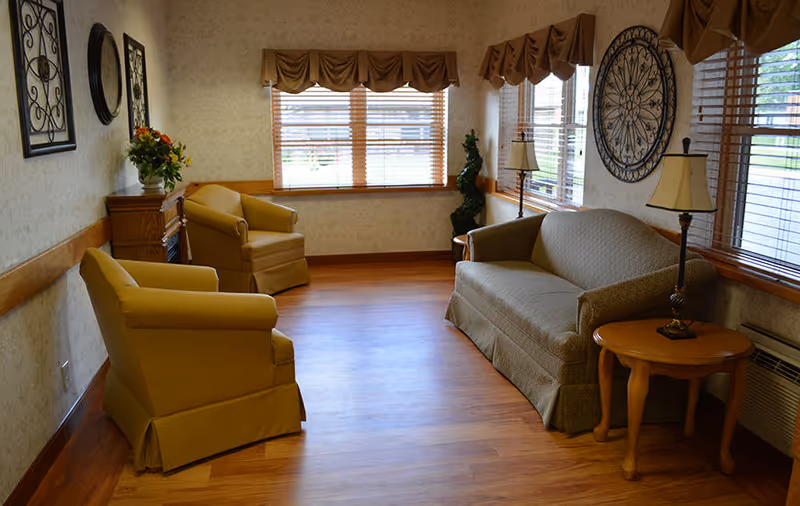 A cozy living room area with wooden flooring, two yellow armchairs, a green sofa, a small wooden side table with a lamp, decorative wall art, and windows with wooden blinds letting in natural light.