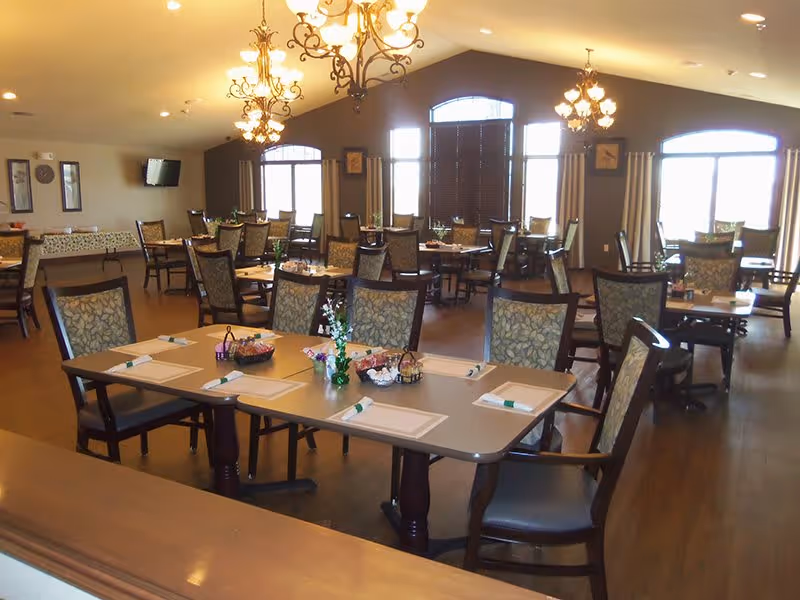 A spacious dining room with multiple tables and chairs arranged neatly. Each table is set with placemats, napkins, and small decorative centerpieces. The room features large windows with curtains, warm lighting from chandeliers, and a wall-mounted television in the background.