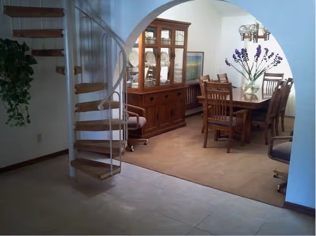 Interior view of a senior living facility showing a dining area with a wooden dining table and six chairs, a glass-front wooden cabinet displaying plates, a vase with purple flowers on the table, a spiral staircase with wooden steps and white railing, and a potted plant hanging on the left wall.