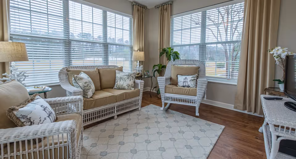 A bright living room with large windows covered by white blinds and beige curtains. The room features white wicker furniture with beige cushions, including a loveseat, an armchair, and a side table with a lamp. There is a patterned area rug on the wooden floor and a TV on a white wicker stand with a remote control on top. A green plant and a white orchid add decorative touches.
