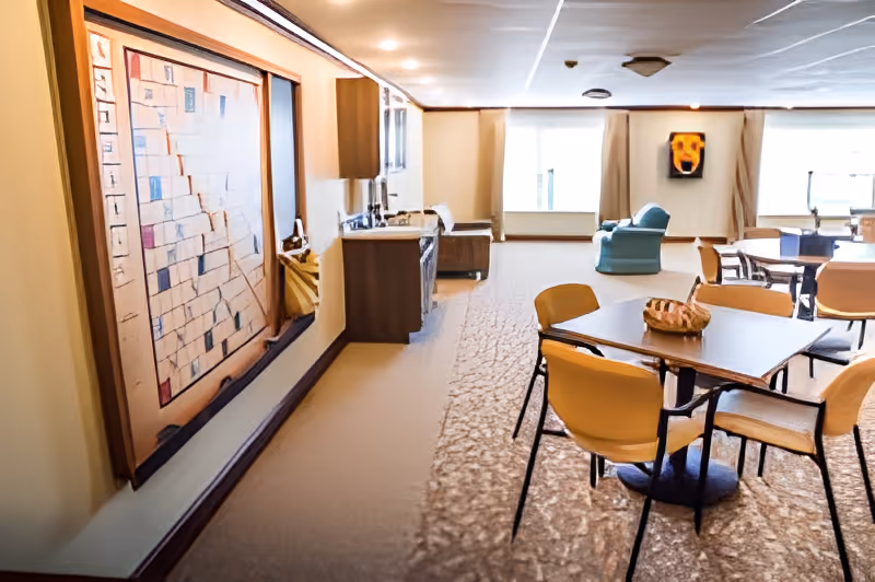 Interior view of a senior living facility common area with tables and chairs arranged for dining or social activities. The room features a carpeted floor, a large framed artwork on the left wall, a small kitchenette area with a sink and cabinets, and a seating area with armchairs near large windows letting in natural light.