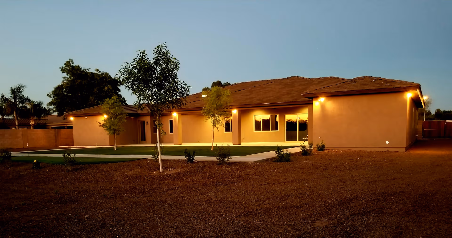 Exterior view of a single-story building at dusk with warm lights illuminating the walls. The building is surrounded by a dirt yard with a few small trees and a concrete pathway leading to the entrance.