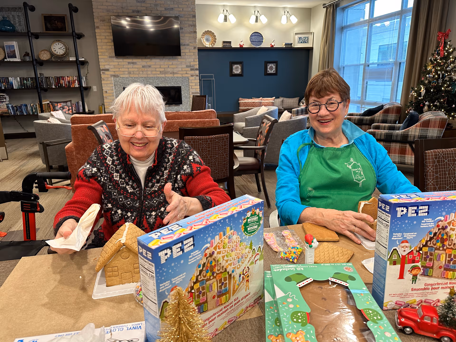 Two elderly women sitting at a table in a cozy living room decorated for Christmas, assembling gingerbread houses with candy decorations. One woman is wearing a red and black patterned sweater and glasses, while the other is wearing a blue jacket and green apron. Behind them are comfortable chairs, a Christmas tree, and a TV mounted on a brick wall.