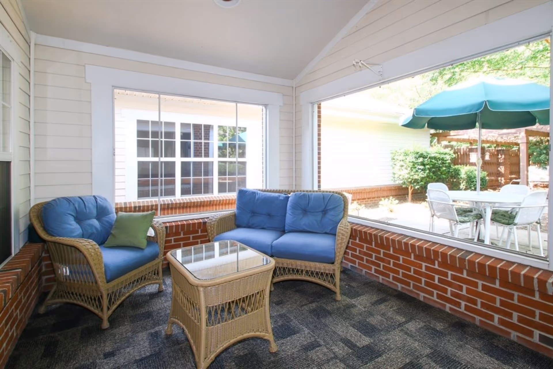 A cozy indoor seating area with wicker furniture including a chair with blue cushions and a green pillow, a loveseat with blue cushions, and a glass-top wicker table. Large windows provide a view of an outdoor patio with a table, chairs, and a green umbrella surrounded by greenery.