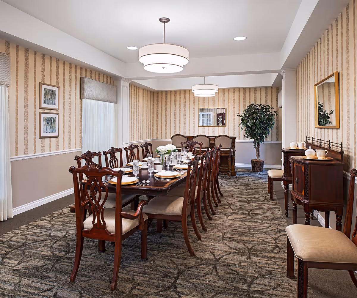 A formal dining room with a long wooden dining table set for a meal with plates, glasses, and silverware. The room features patterned wallpaper, carpeted floor, two large ceiling lights, framed pictures on the wall, a potted plant, and wooden sideboards with decorative items.