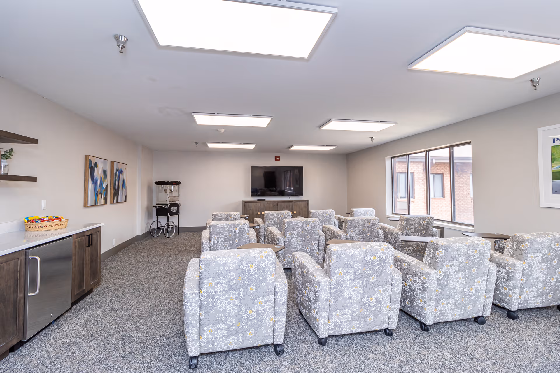 Community media room with rows of patterned armchairs facing a wall-mounted TV, a kitchenette on the left, and large windows on the right.