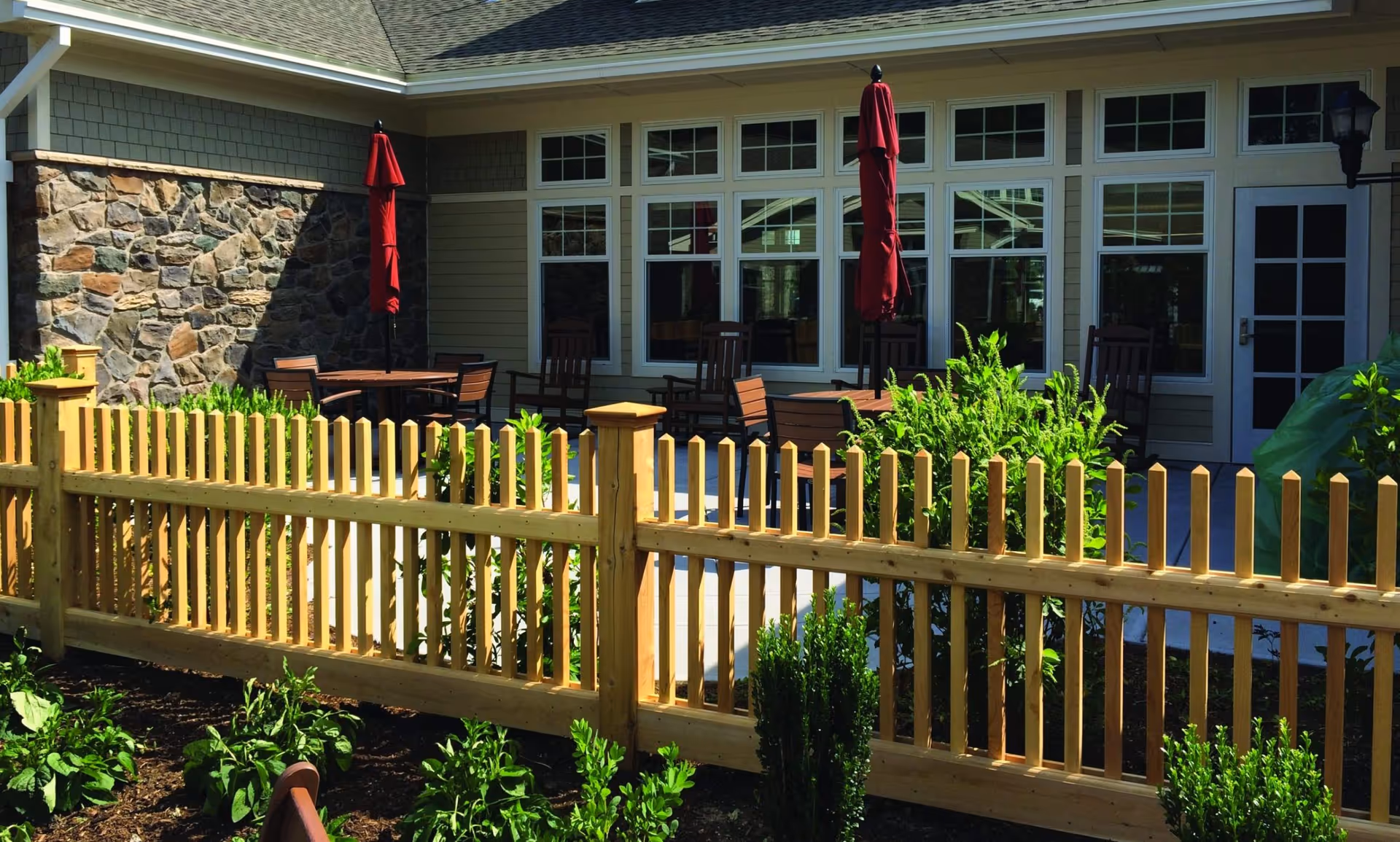 Outdoor patio area with wooden tables and chairs, two closed red umbrellas, a wooden picket fence, green plants, and a building with stone and siding walls and multiple windows in the background.