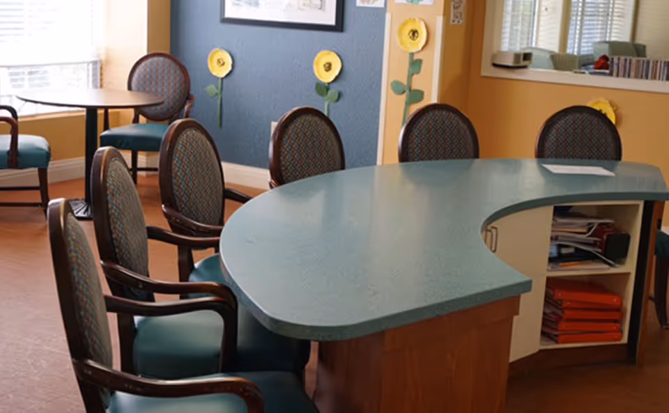 Interior view of a senior living facility common area with a curved reception desk and several upholstered chairs arranged around it. The walls are decorated with colorful paper flowers and framed artwork. There is a window letting in natural light and additional seating with small round tables in the background.