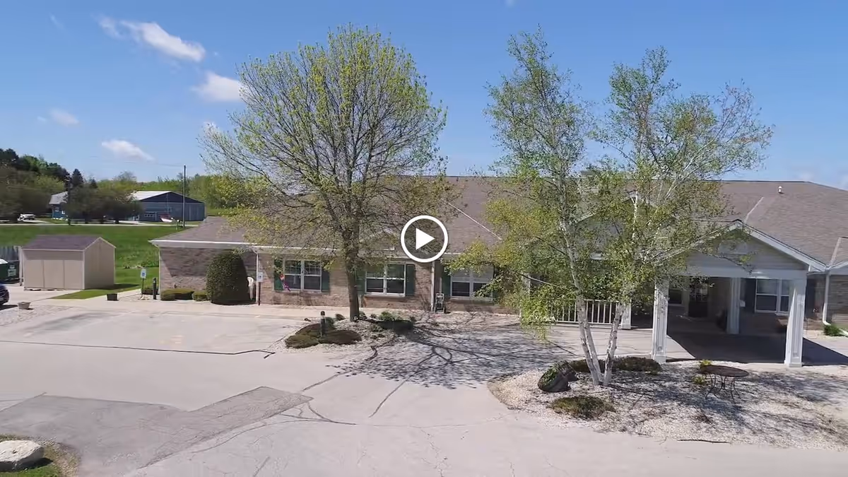 Exterior view of Meadow View Assisted Living facility showing a single-story brick building with a covered entrance, surrounded by trees and a paved parking area under a clear blue sky.