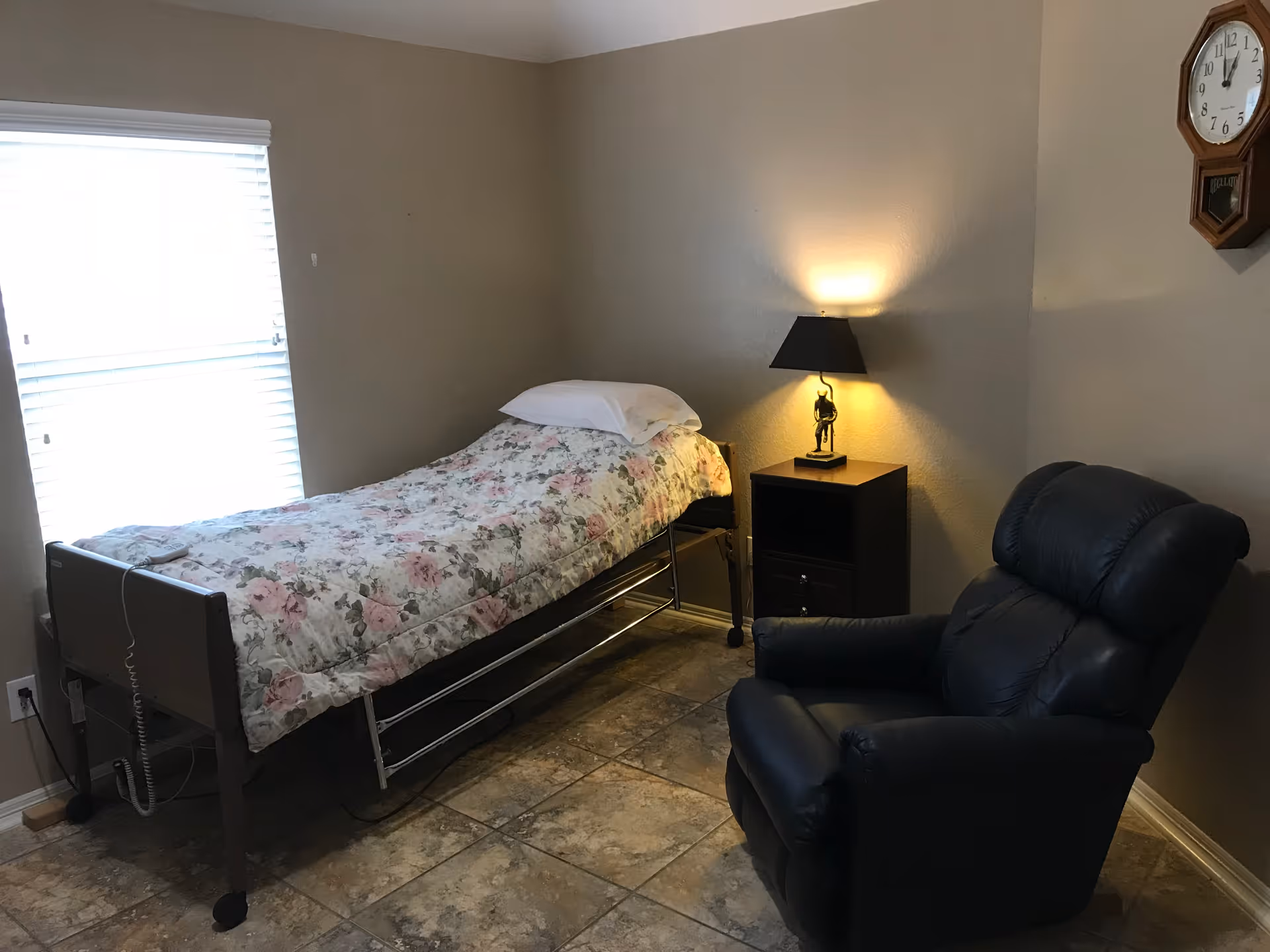 A small bedroom with a single bed covered with a floral bedspread, a white pillow, a bedside table with a lamp, a black recliner chair, a window with blinds, and a wall clock.