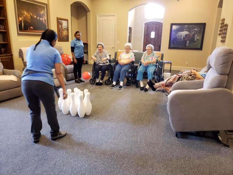 A group of elderly women, some in wheelchairs, are seated in a living room area watching a staff member roll a ball towards a set of bowling pins on the carpet. Another staff member stands nearby observing. The room has comfortable chairs, framed pictures on the walls, and a door with a window letting in natural light.