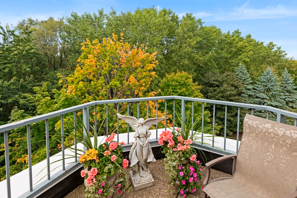 A balcony with a metal railing overlooking a lush green landscape with trees, some showing early autumn colors. The balcony has a beige cushioned chair, a stone angel statue, and two planters filled with pink and yellow flowers and green foliage.