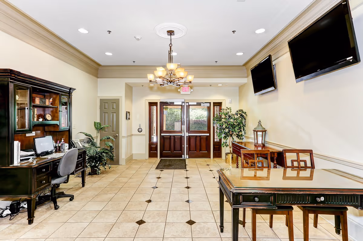 Interior view of a well-lit room with tiled floor, featuring a dark wooden desk with a computer and chair on the left, a glass-top table with chairs on the right, two wall-mounted flat-screen TVs, potted plants, and double wooden doors with glass panels at the far end.
