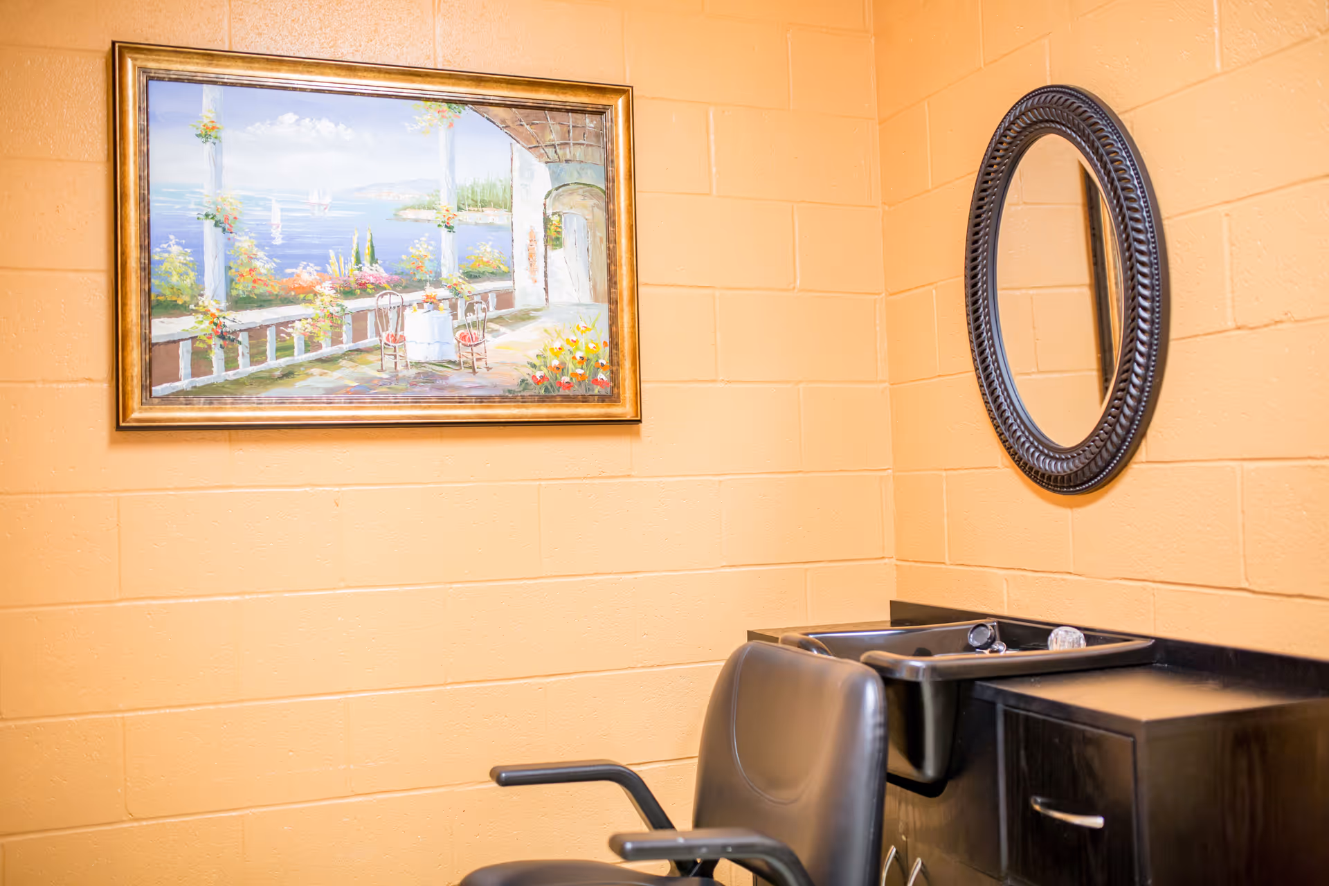 Interior view of a small room with peach-colored brick walls, featuring a black salon chair in front of a black sink and cabinet. A round mirror with a decorative frame hangs on the wall above the sink, and a framed painting of an outdoor terrace overlooking a body of water with flowers and chairs is mounted on the adjacent wall.