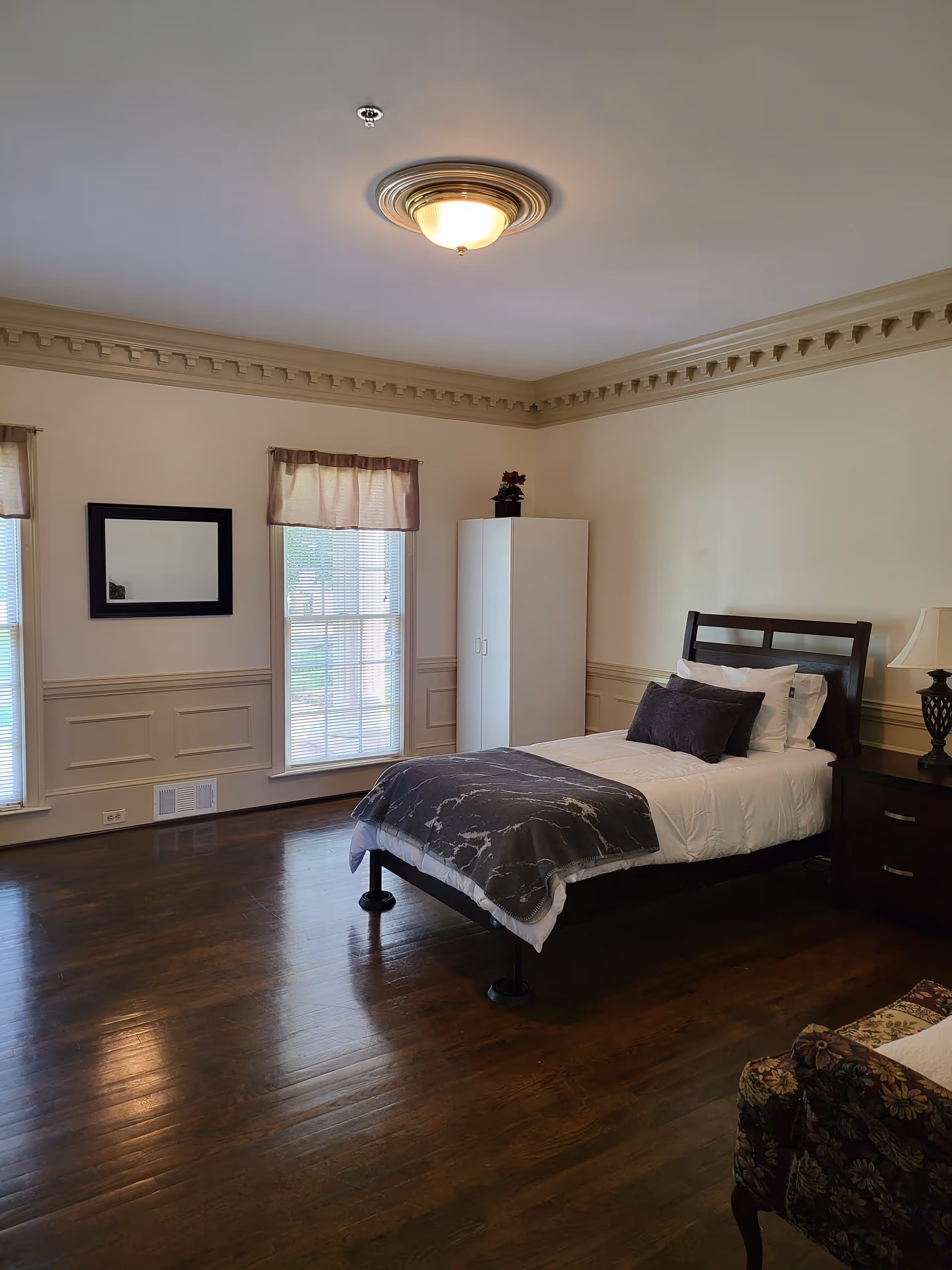 A neatly made single bed with white and dark gray bedding in a spacious room with wooden floors. The room has two windows with light curtains, a white wardrobe, a dark wooden nightstand with a lamp, a wall mirror, and a floral upholstered chair partially visible in the corner.