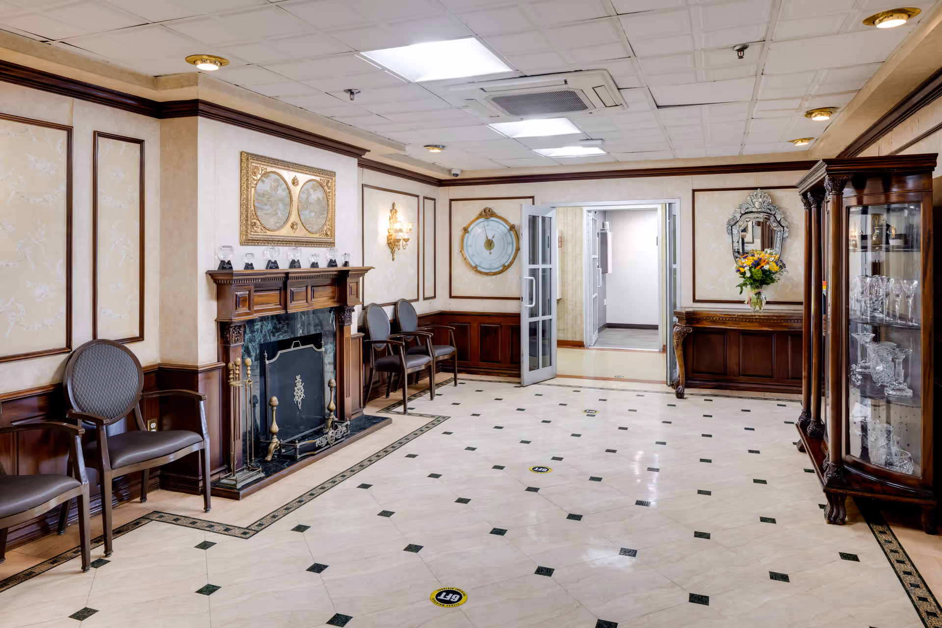 An elegant waiting area in a nursing home featuring a decorative fireplace with a dark wood mantle, several chairs along the walls, a glass display cabinet with crystal items, a wall clock, a mirror above a wooden console table with a vase of flowers, and a tiled floor with social distancing markers.