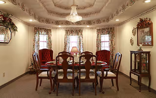 A formal dining room with a wooden dining table surrounded by six chairs, two of which have red upholstery. The room features ornate ceiling molding, a chandelier, floral curtains on three windows, a lamp on the table, a small display cabinet, and decorative wall hangings.