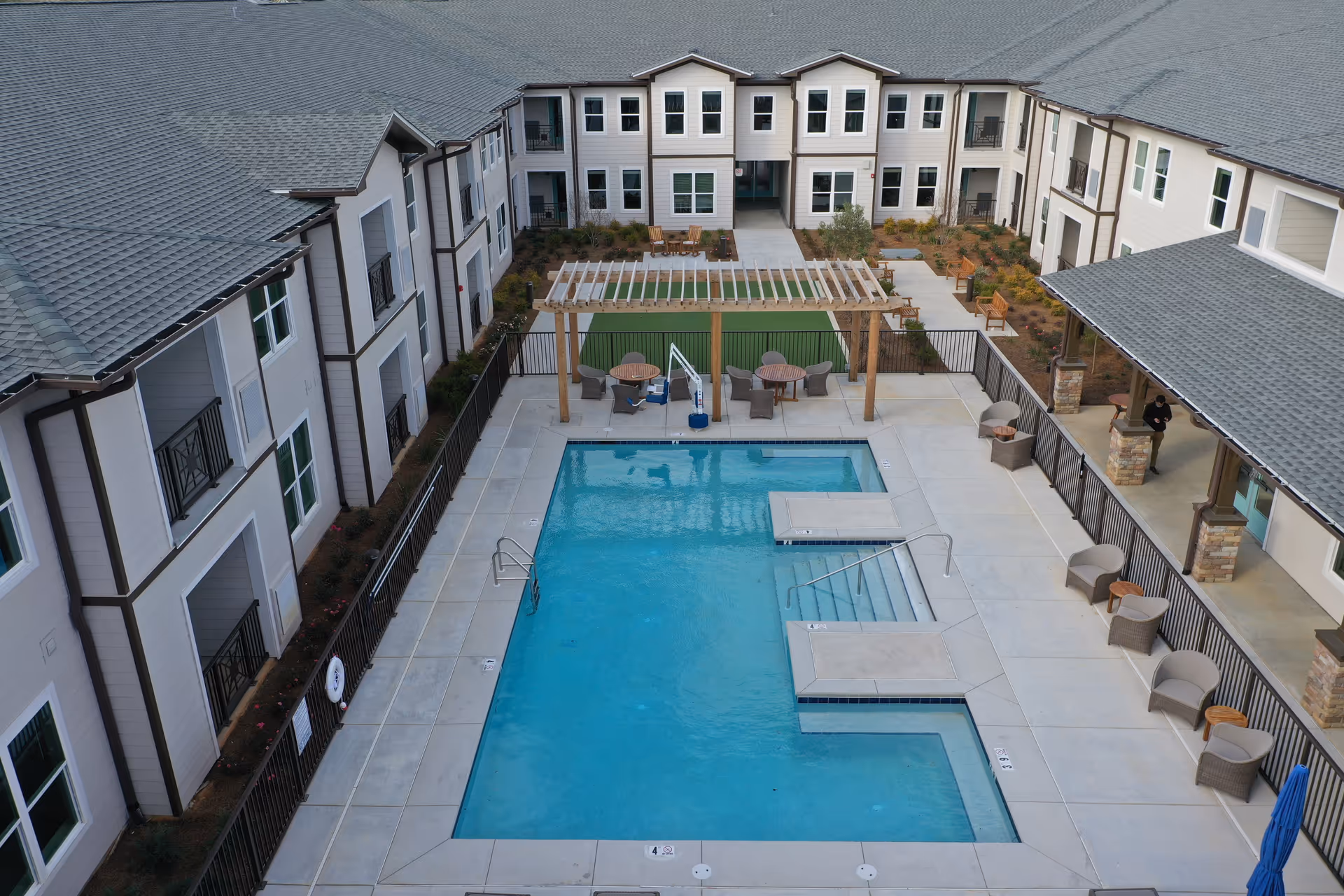 Aerial view of an outdoor swimming pool area surrounded by a multi-story residential building. The pool area features lounge chairs, tables with umbrellas, a pergola with seating underneath, and landscaped garden beds. The building has multiple windows and balconies overlooking the pool.