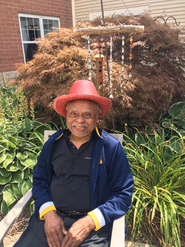 An elderly man wearing a red hat and a navy blue jacket with yellow trim is sitting on a chair outdoors in a garden area with green plants and a bush behind him. A brick building and a window are visible in the background.