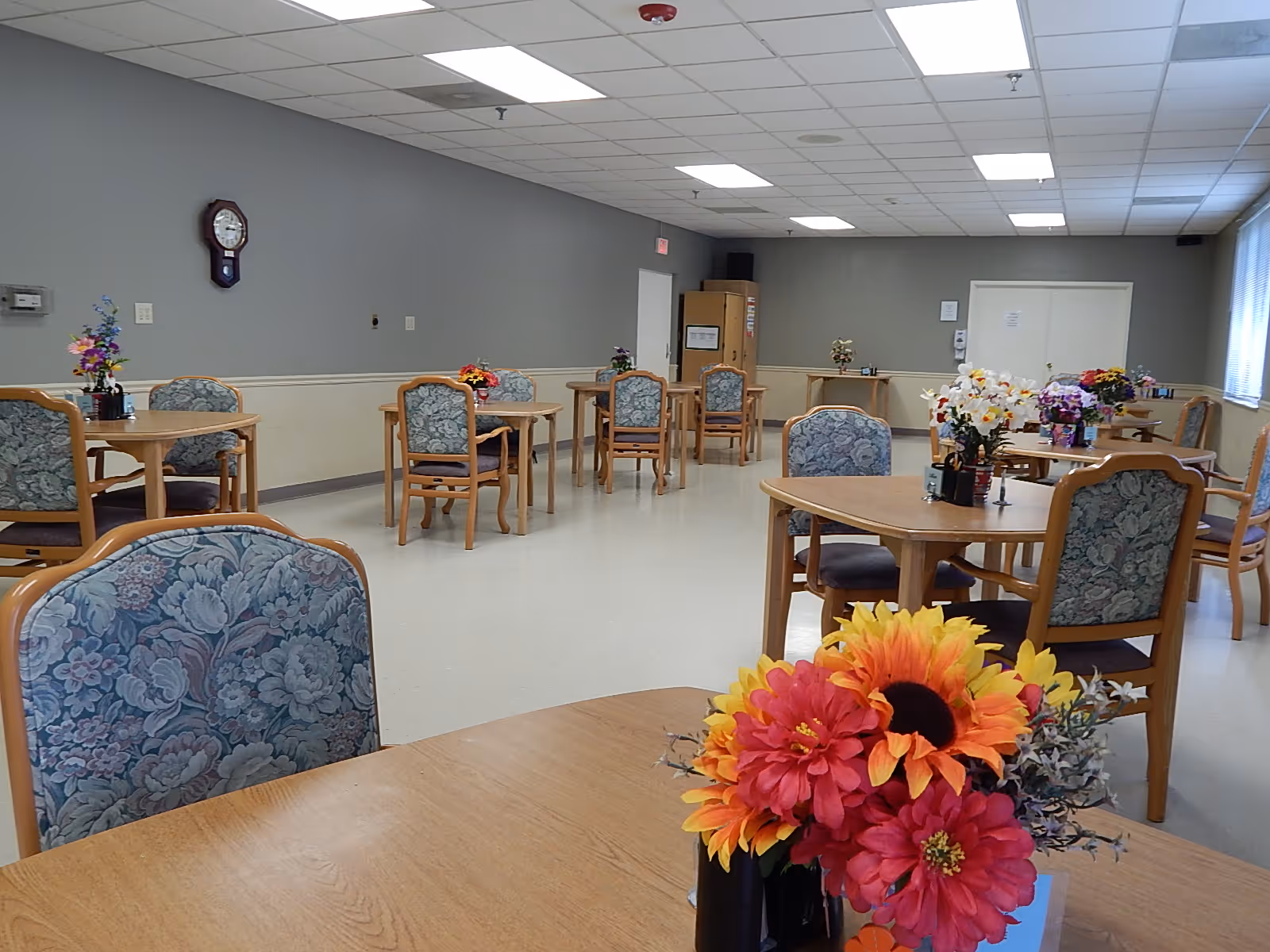 A dining room with multiple wooden tables and chairs arranged neatly. Each table has a floral centerpiece with colorful flowers. The room has gray walls, a clock on one wall, and a ceiling with fluorescent lights. There are windows on the right side letting in natural light.