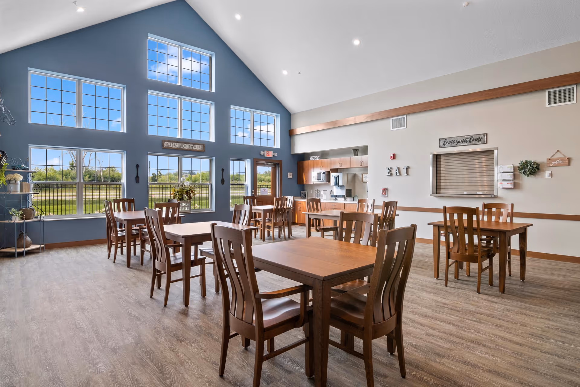 A bright dining room with multiple wooden tables and chairs arranged neatly. Large windows with a blue accent wall let in natural light and offer a view of greenery outside. The room features a high vaulted ceiling with recessed lighting. On one wall, there is a serving window with a sign above it that reads 'Come sweet home' and decorative letters spelling 'EAT'.