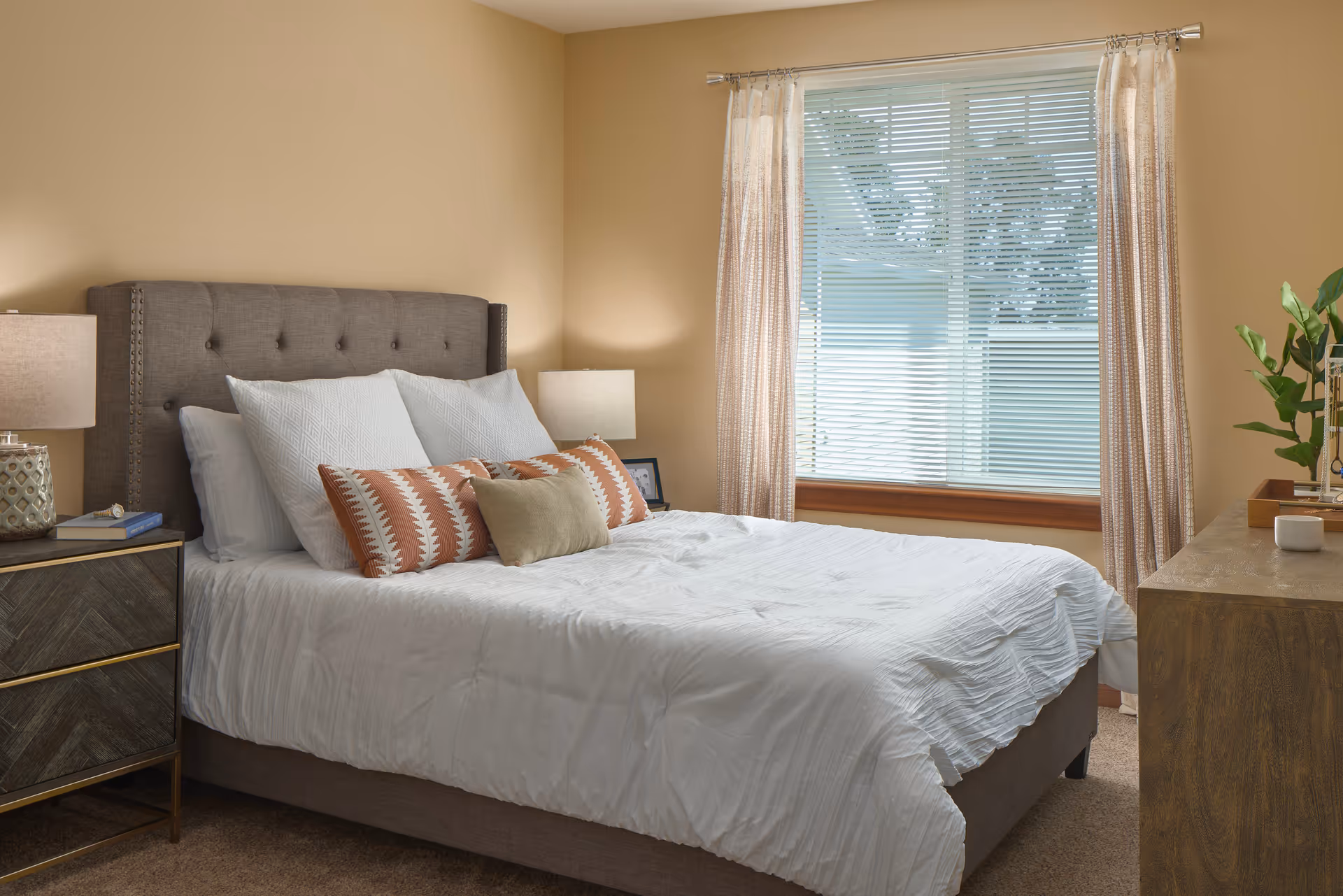 A neatly made bed with a tufted gray headboard, white bedding, and decorative pillows in white, orange, and beige. There are two bedside tables with lamps on either side of the bed. A large window with blinds and light-colored curtains is on the right wall, and a wooden dresser with a plant and decorative items is visible on the right side of the room.