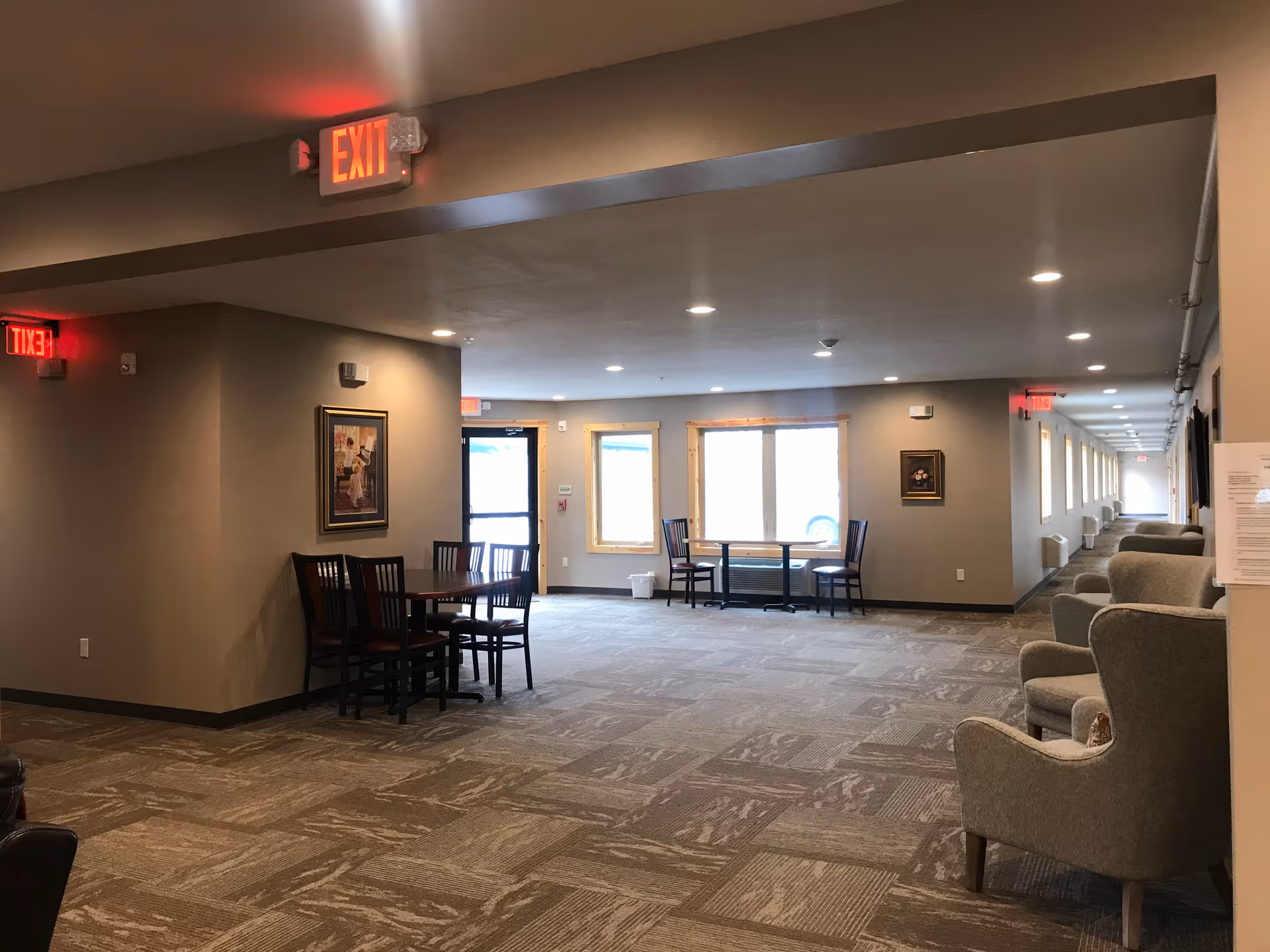 Spacious interior common area with seating and tables, large windows, carpeted floor, and a long hallway with illuminated exit signs.