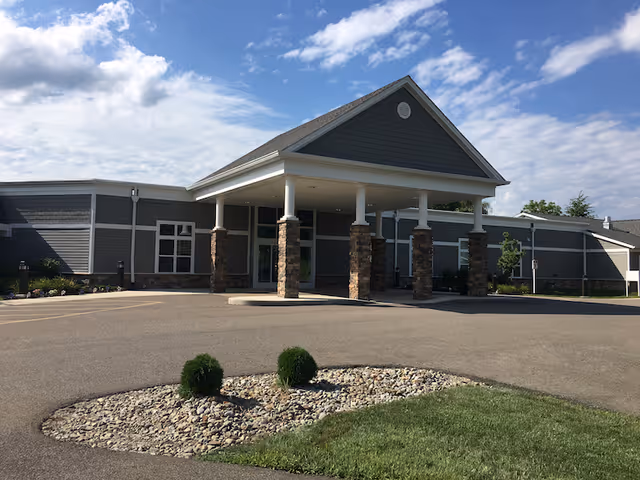 Exterior view of a single-story healthcare facility building with a covered entrance supported by stone pillars. The building has gray siding with white trim and a landscaped area with small bushes and rocks in front. The sky is partly cloudy.