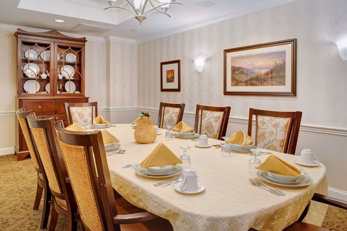A formal dining room with a long table covered by a cream-colored tablecloth, set with plates, cups, silverware, and folded yellow napkins. The room features wooden chairs with floral upholstery, a wooden china cabinet displaying plates and teapots, patterned wallpaper, and framed artwork on the walls.