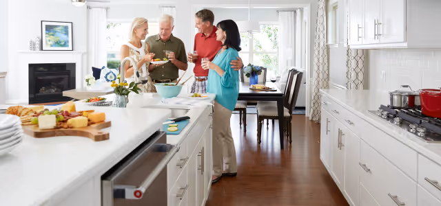 Four adults standing in a bright, modern kitchen with white cabinets and a wooden floor. They are smiling and holding drinks, engaging in conversation near a kitchen island with food and plates on it. A dining table with chairs is visible in the background near large windows with white curtains.