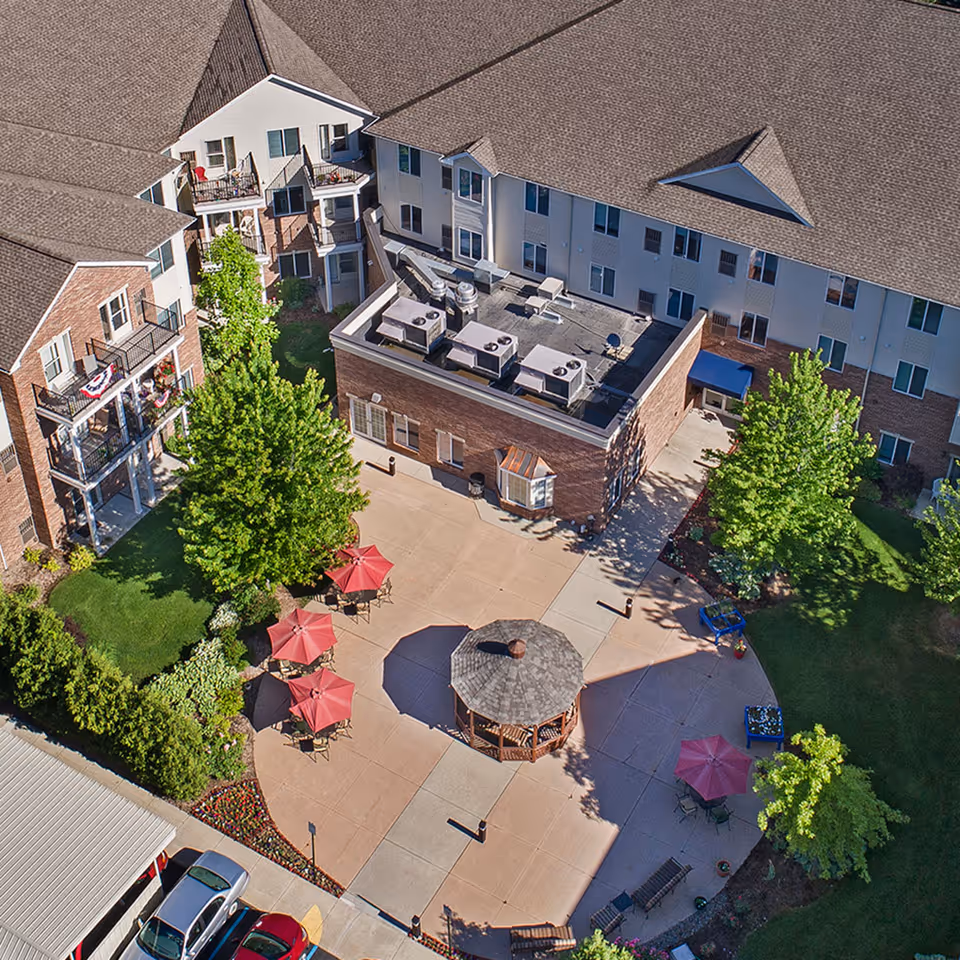 Aerial view of an outdoor courtyard area at American House Sterling Woods featuring a central gazebo, several red umbrellas with tables and chairs, surrounded by trees and a multi-story residential building with balconies.