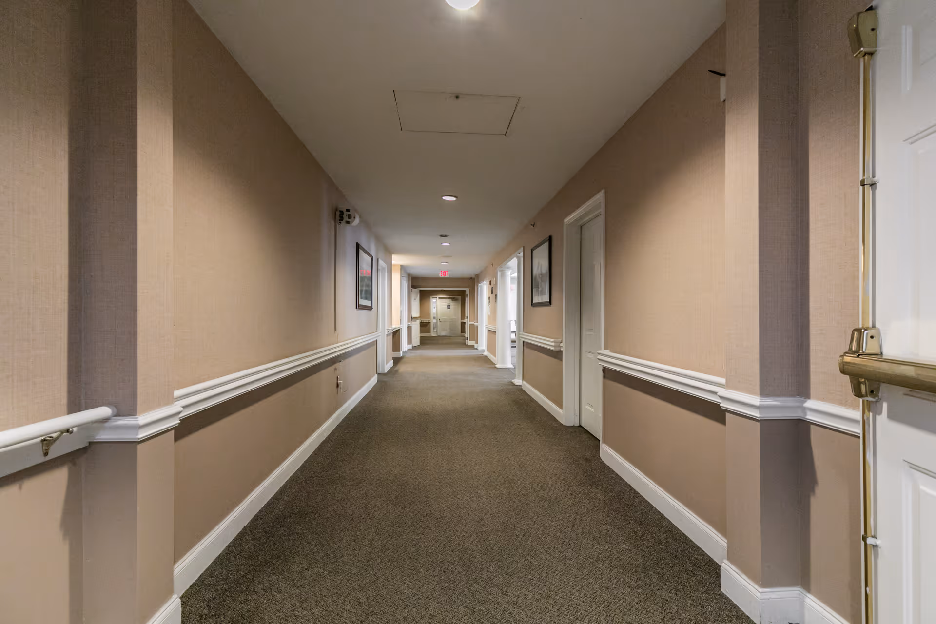 A long, carpeted hallway inside a senior living facility with beige walls, white trim, handrails on both sides, framed pictures on the walls, and several closed doors along the corridor. The hallway is well-lit with ceiling lights and has an exit sign visible at the far end.