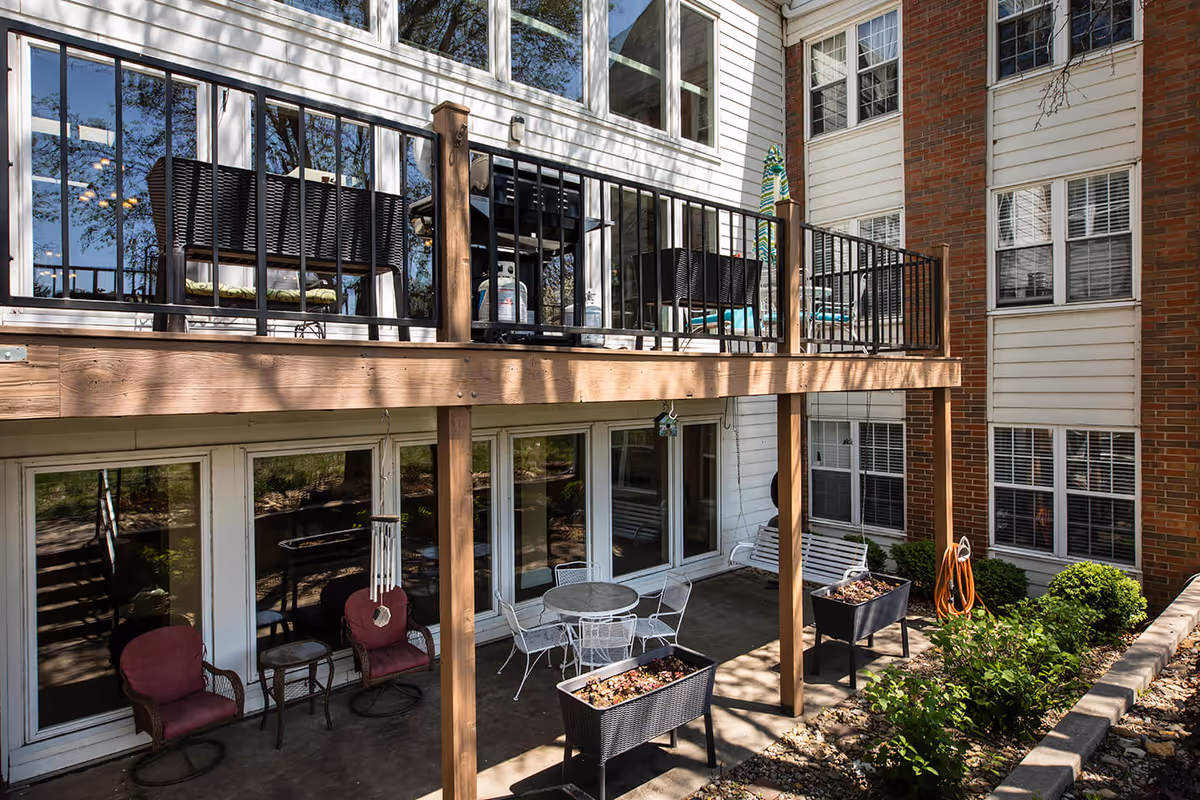 Outdoor patio area of Lexington Park Assisted Living of Topeka featuring a wooden deck with black metal railing, outdoor seating including chairs with cushions, a round table with chairs, a white porch swing, and planter boxes with plants. The building exterior has white siding and brick walls with multiple windows.