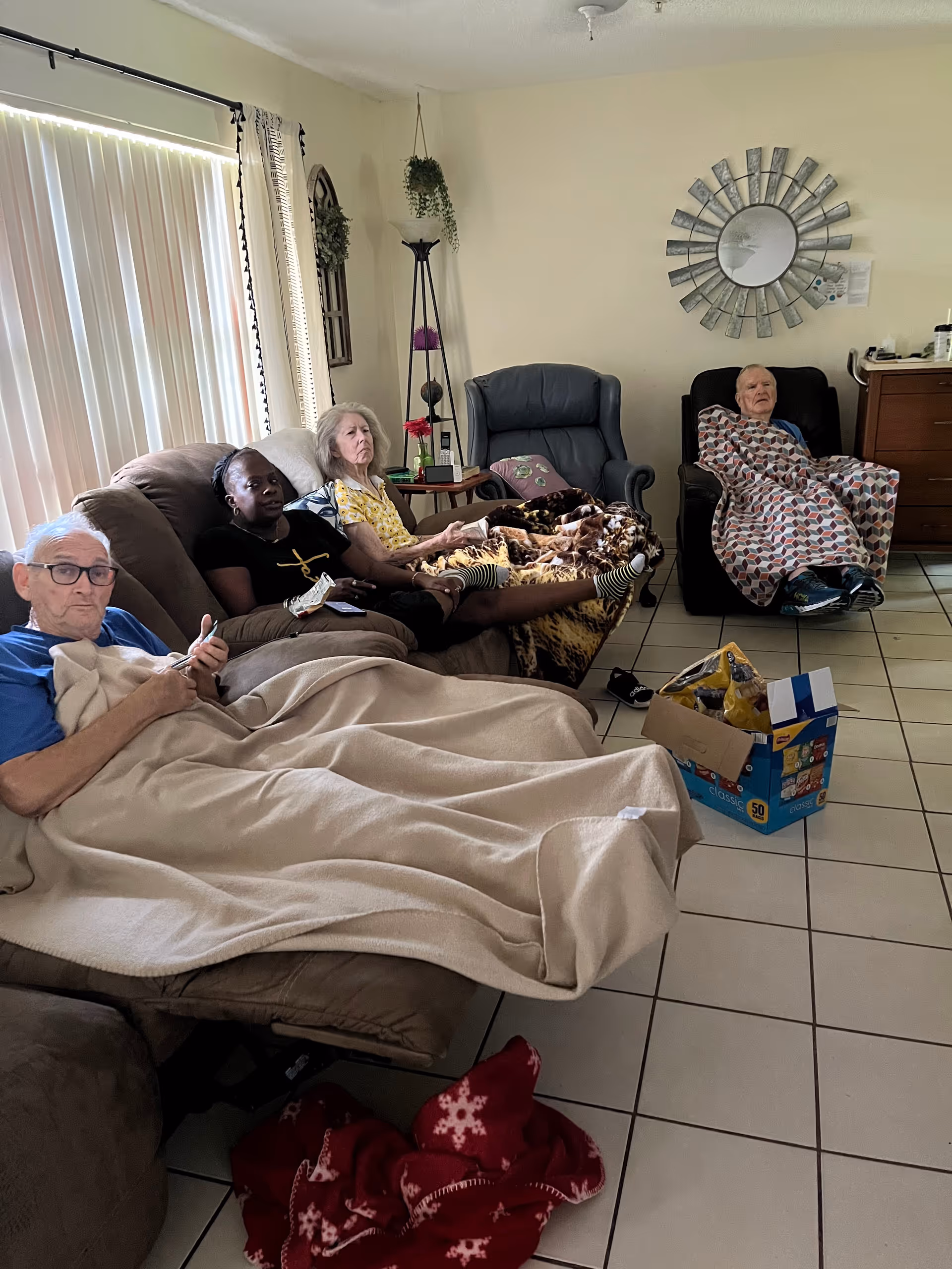 Four elderly individuals sitting in a living room area. Three are seated on a couch covered with blankets, and one is in a recliner chair also covered with a blanket. The room has tiled floors, a large window with vertical blinds, a decorative mirror on the wall, a small table with a lamp and plants, and a box of snacks on the floor.