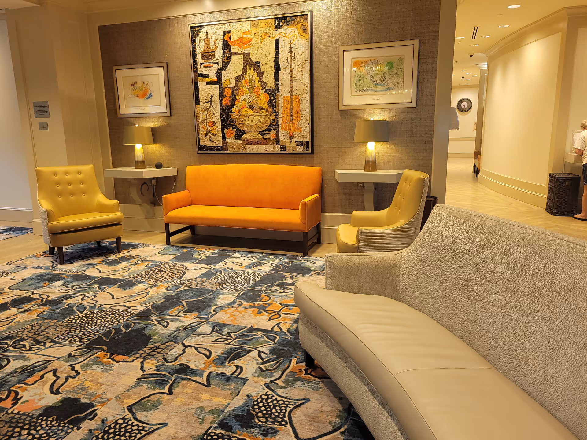 A cozy seating area in a senior living facility with a yellow sofa, two yellow armchairs, and a beige couch arranged on a patterned carpet. The wall behind the yellow sofa features two framed artworks and two wall-mounted lamps on small white shelves. A hallway is visible to the right with a person standing near a trash bin.