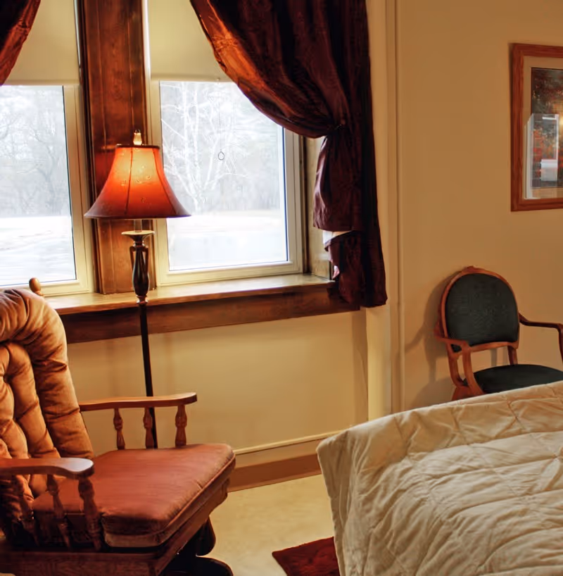 A cozy bedroom corner with a cushioned wooden rocking chair, a floor lamp with a red lampshade, two windows with dark red curtains, a green cushioned wooden chair, and a bed with a white quilted blanket.