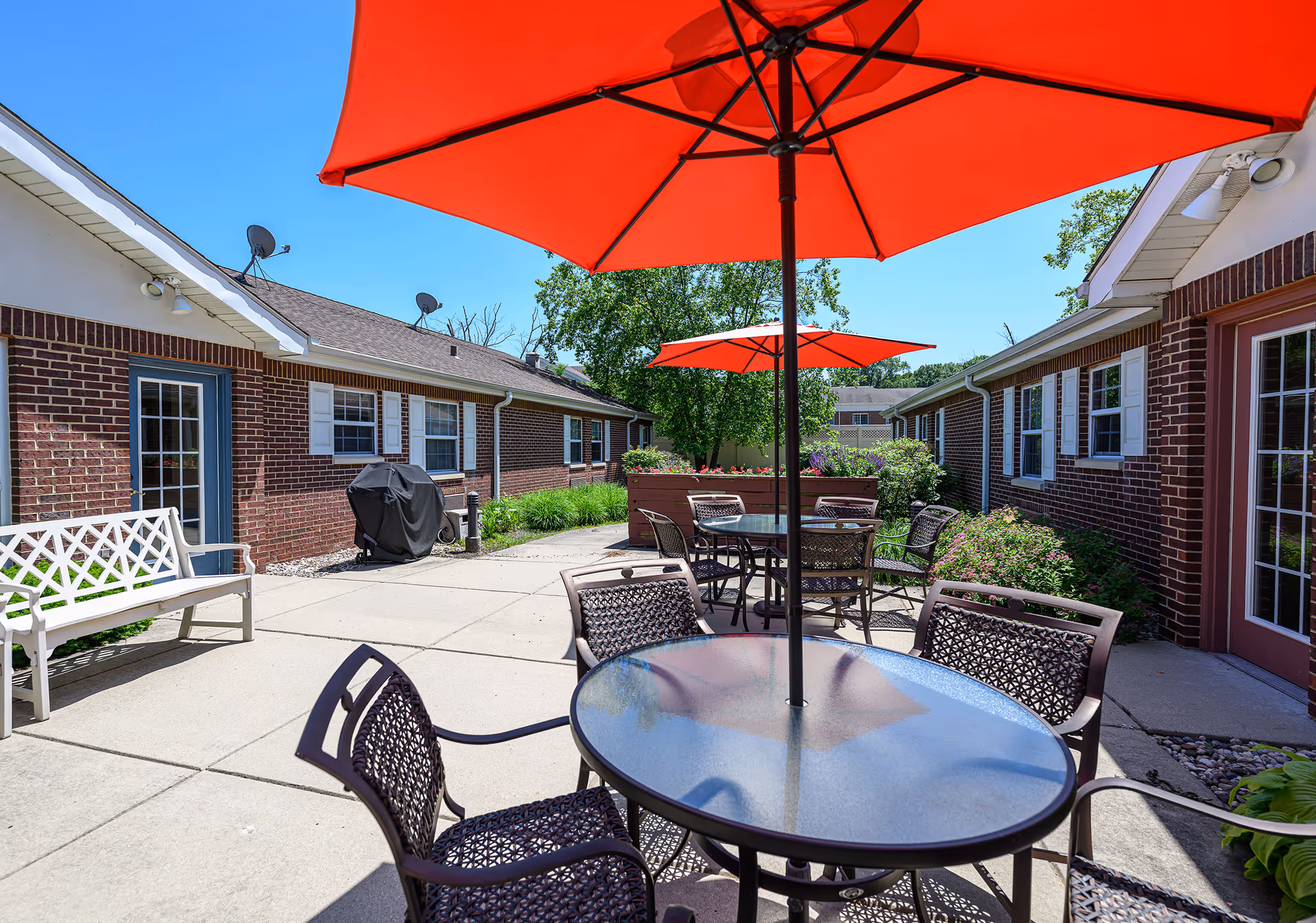 Outdoor patio area with round glass tables and metal chairs under large red umbrellas. The patio is surrounded by single-story brick buildings with white-framed windows and doors. There is a white bench and a covered grill on the left side, with greenery and flower beds along the edges. The sky is clear and blue.