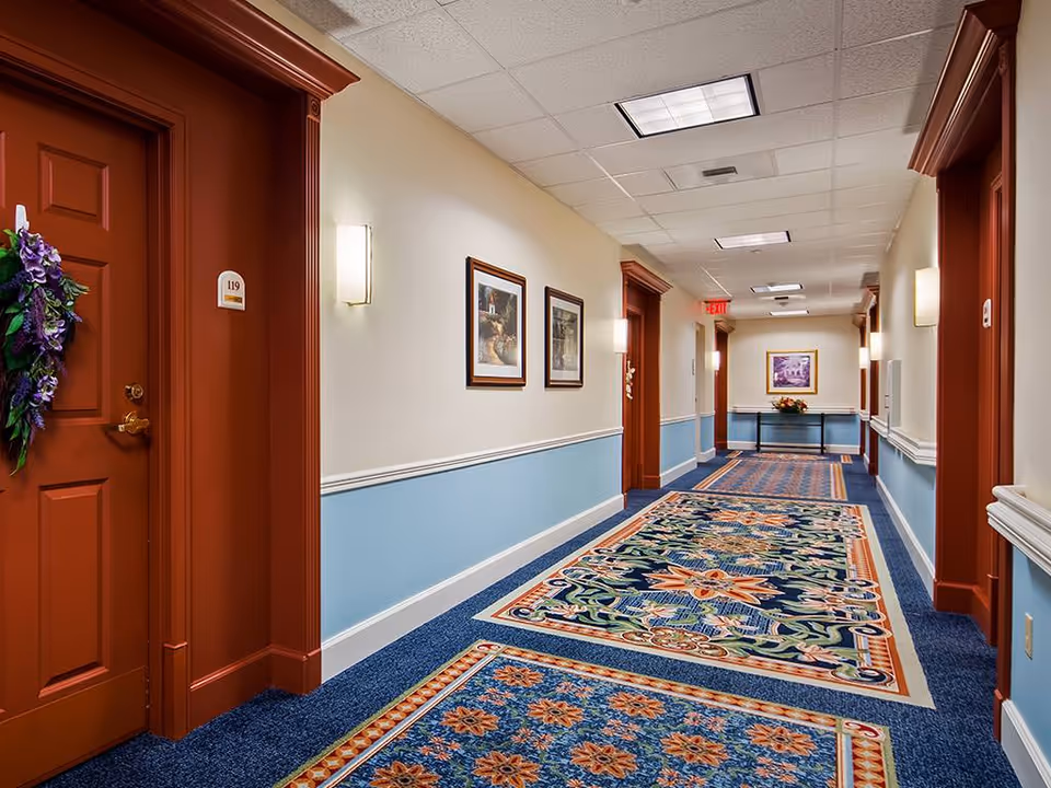 Carpeted interior hallway with patterned runner rugs, framed pictures, wall sconces, and red-painted apartment doors.
