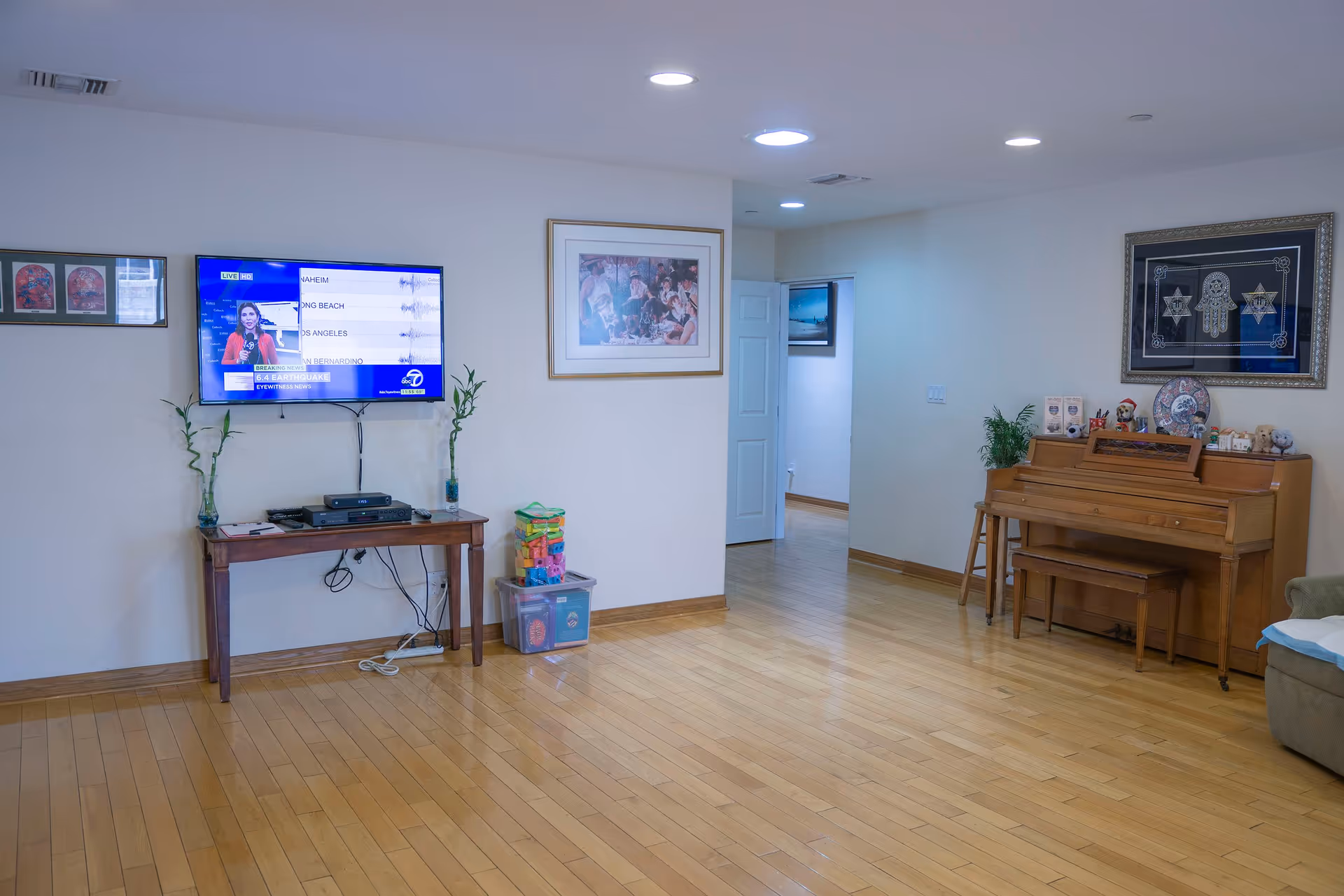 A spacious living room with light wooden flooring and white walls. On the left wall, a flat-screen TV is mounted above a small wooden table with two bamboo plants and electronic devices. Next to the table are two plastic storage bins with colorful toys. On the right side, there is a wooden piano with a matching bench, decorated with various small items and a framed artwork above it. The room is well-lit with recessed ceiling lights and has an open doorway leading to another room.