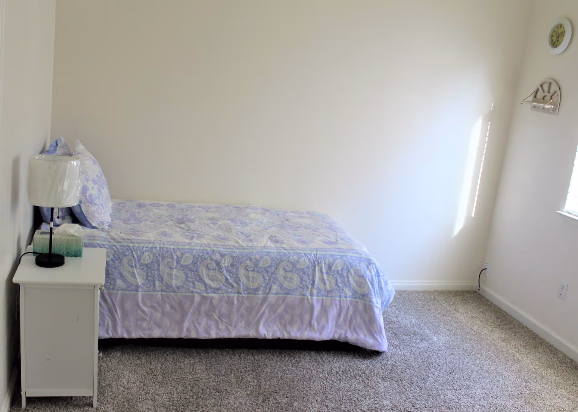 A simple bedroom with a single bed covered in a purple patterned bedspread, a white nightstand with a lamp and tissue box, beige carpet, and a clock on the wall near a window letting in natural light.