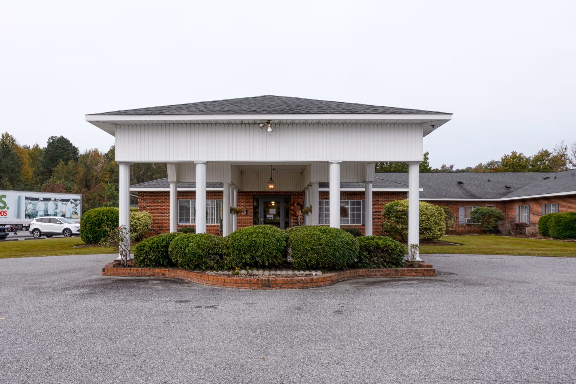 Front exterior view of a single-story brick building with a covered entrance supported by white columns, surrounded by neatly trimmed bushes and a paved driveway.