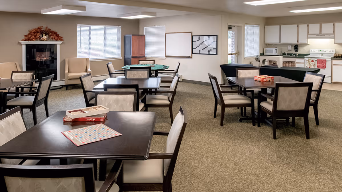 A spacious common room with several tables and chairs arranged for group activities. On one table, there is a Scrabble game set up, and on another table, a Yahtzee game box is visible. The room has beige walls, carpeted floors, a fireplace with autumn decorations, and a small kitchen area with white cabinets and appliances in the background.